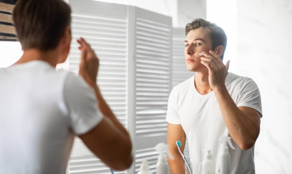 Male Facial Skincare Concept. Portrait Of Confident Handsome Man Applying Moisturizing Cream On Smooth Face Caring For Skin After Shaving Routine, Looking In The Mirror, Standing In Bathroom At Home