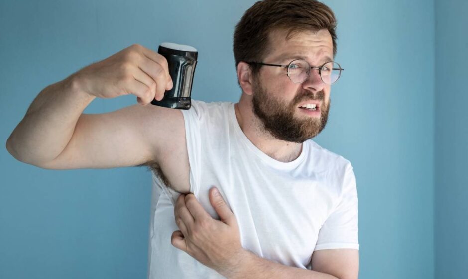 Man is irritated by his sweating and was about to use an antiperspirant to prevent the unpleasant smell of armpit sweat. Isolated, on a blue background.