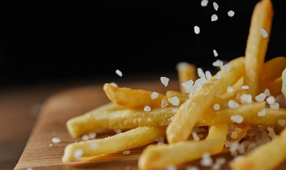 close up of salt falling on fresh golden french fries on wooden chopping board