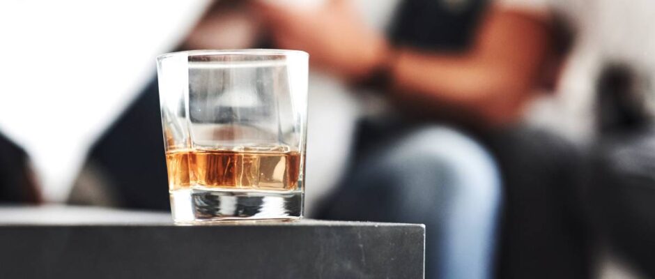 Close up focused photo of glass with whiskey standing on the black table with blurred people on the background.