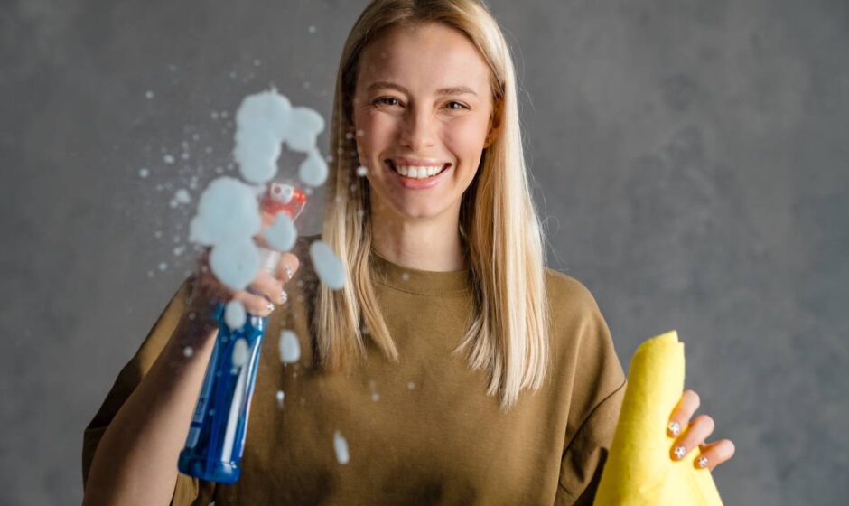 Happy blonde woman maid holding bottle of detergent and a rag, spraying glass